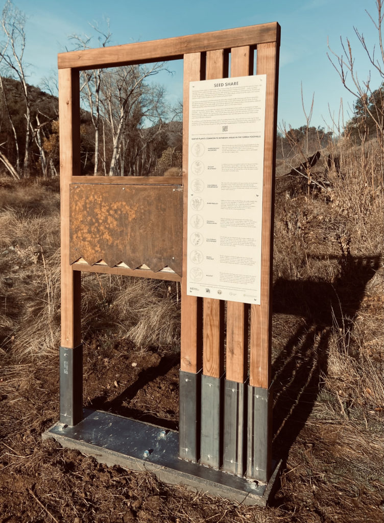 Seed Share: wood kiosk standing in a field. The kiosk is embedded with a seed packet dispenser and has informational signage attached. 
